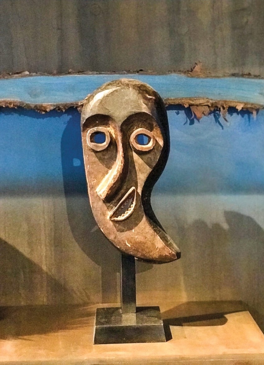 African tribal art sculpture with abstract face, displayed on a stand against a blue and brown textured background.