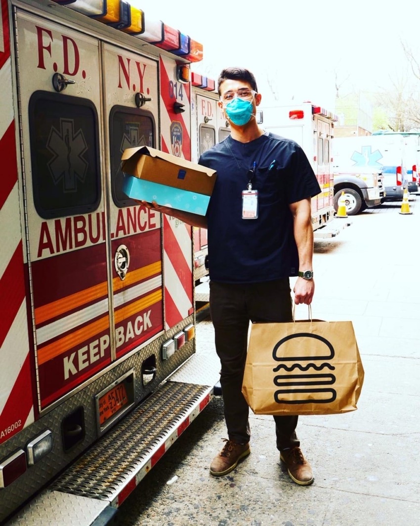 Healthcare worker with mask holding food delivery bags in front of NYC ambulance.