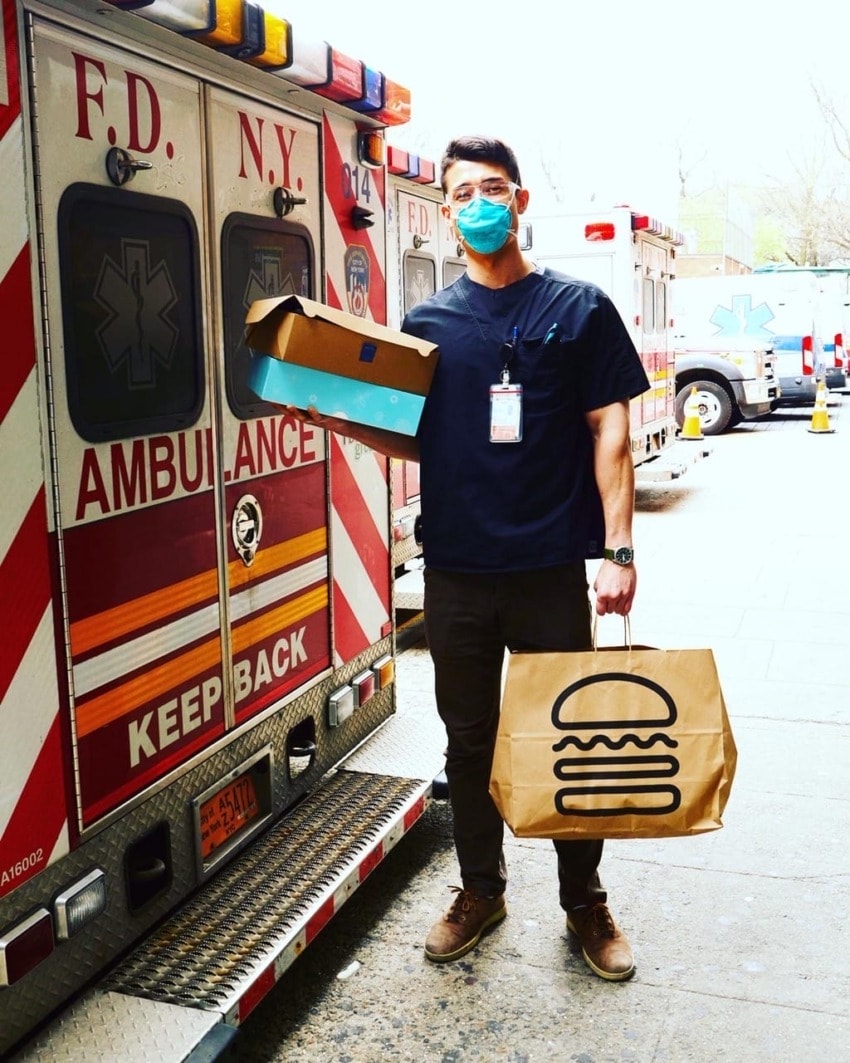 Healthcare worker with mask holding food delivery bags in front of NYC ambulance.