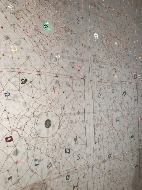 Intricate wire and thread artwork with scattered letters and symbols on a wall, creating a complex geometric design.