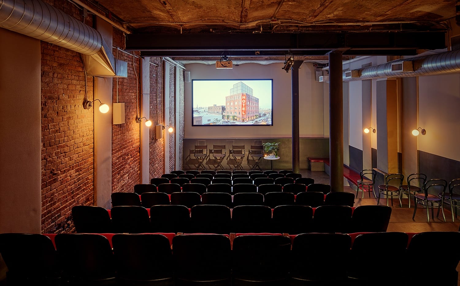 Small theater with red brick walls, empty black chairs, and a large screen displaying an urban building.