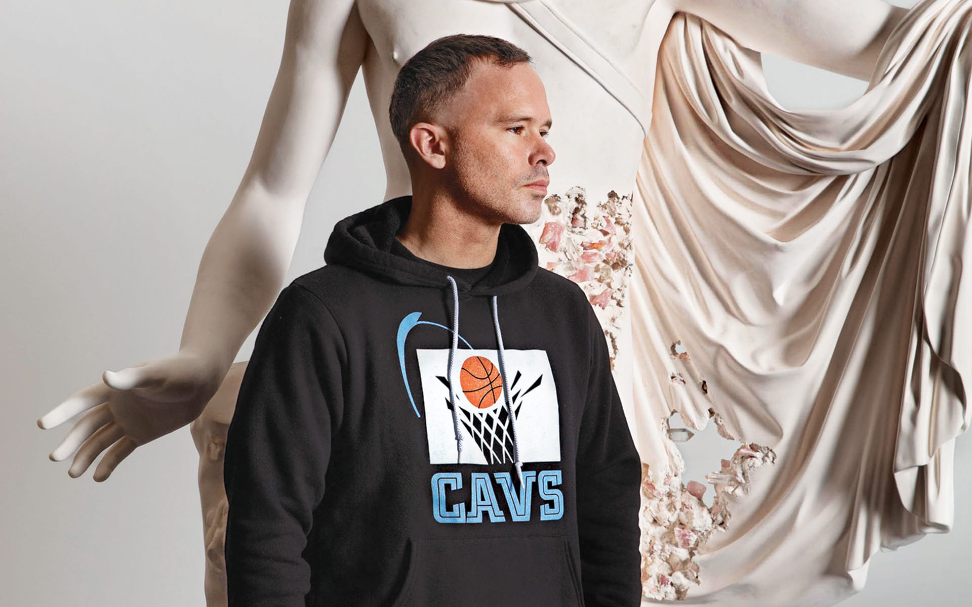 Man in black CAVS hoodie standing in front of a classical statue with draped fabric and floral details.