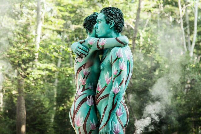 Two people embrace, bodies painted with green and pink floral designs, set against a lush forest background.