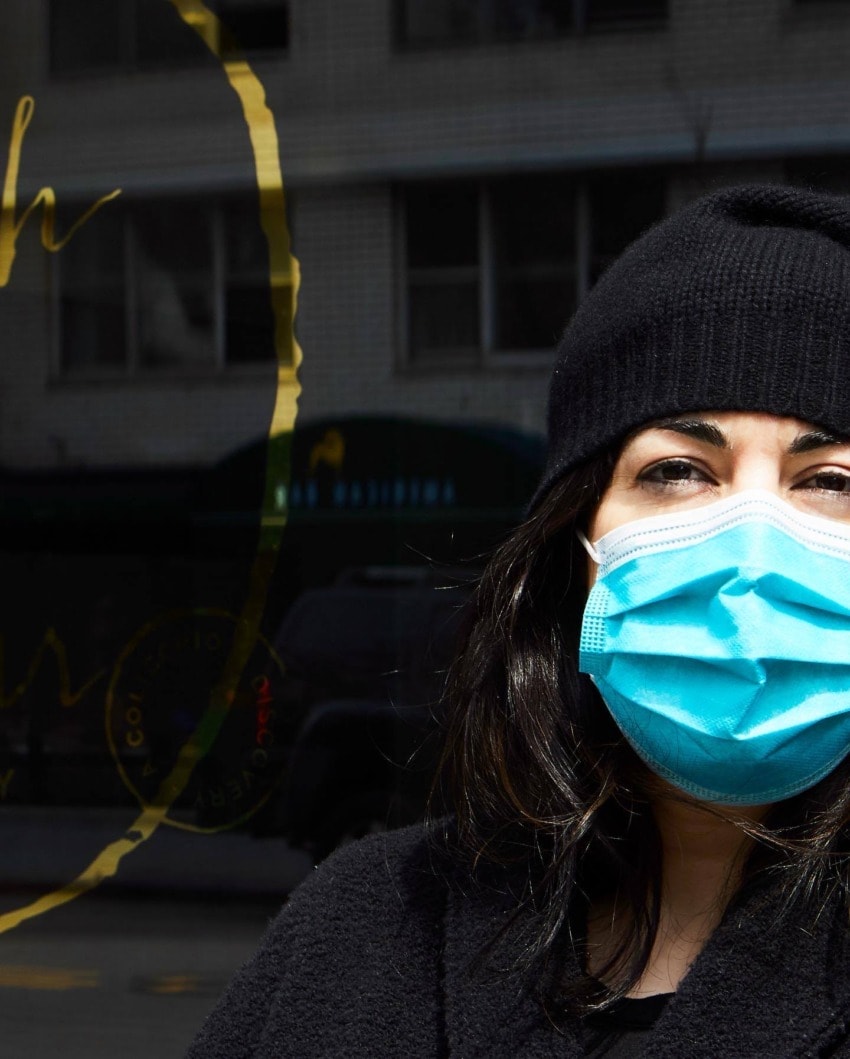 Person wearing a blue face mask and black beanie standing outdoors on a sunny day with building in the background