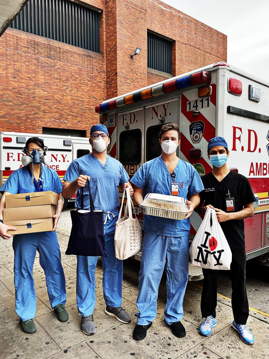 Healthcare workers in scrubs and masks stand by an ambulance holding bags and boxes in a city setting.
