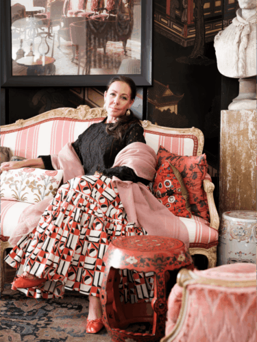 Woman sitting on an ornate sofa in a stylish room with patterned skirt and pink shawl, surrounded by decorative elements.