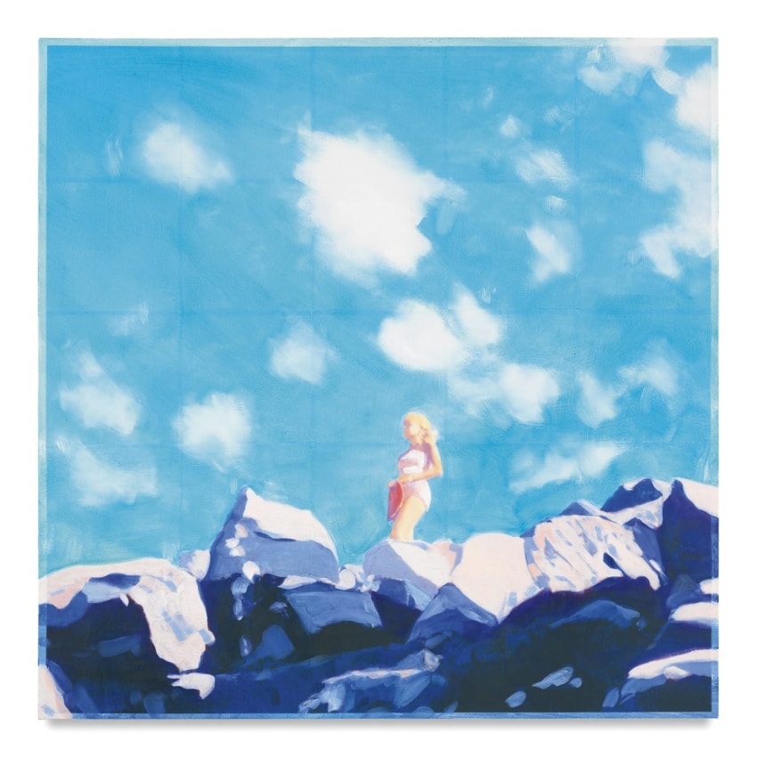 Woman standing on rocks under a bright blue sky with white clouds overhead.