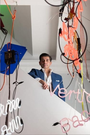 Person in a blue suit stands on stairs surrounded by colorful neon lights and wires.
