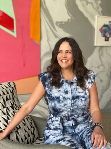 Person sitting on a chair smiling, wearing a blue tie-dye outfit, with colorful abstract art in the background.