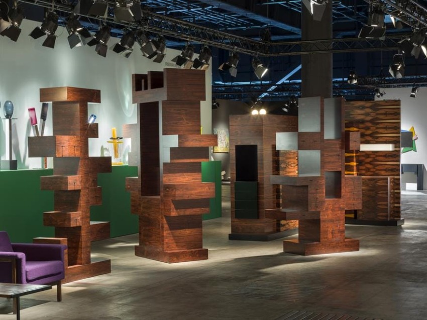 Modern art exhibition with tall, abstract wooden sculptures in a room with spotlights and colorful background objects.