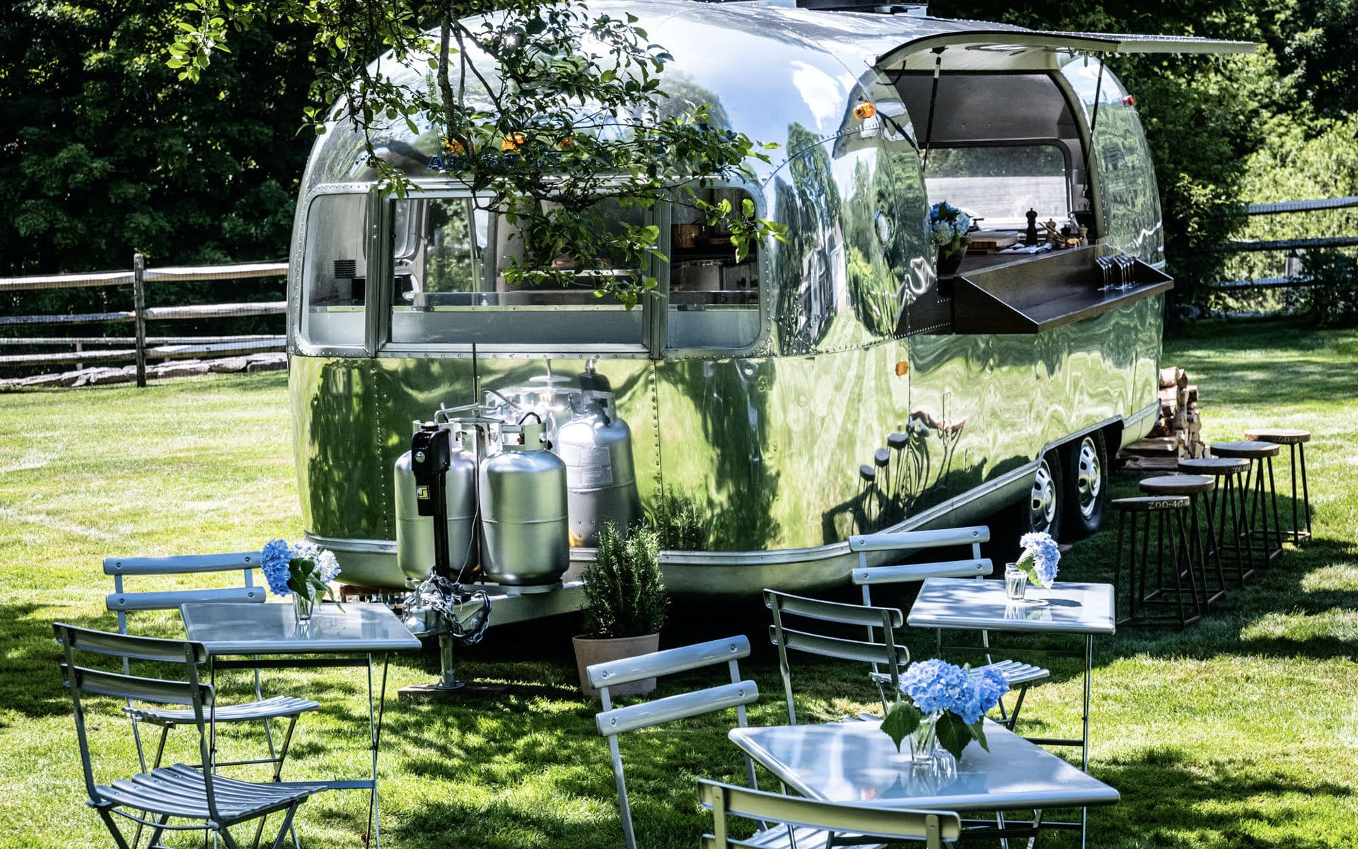 Vintage silver food trailer in a grassy area with blue flowers on tables and wooden fence in the background.