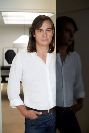 Person with long hair in a white shirt standing indoors next to a reflective surface showing their reflection.