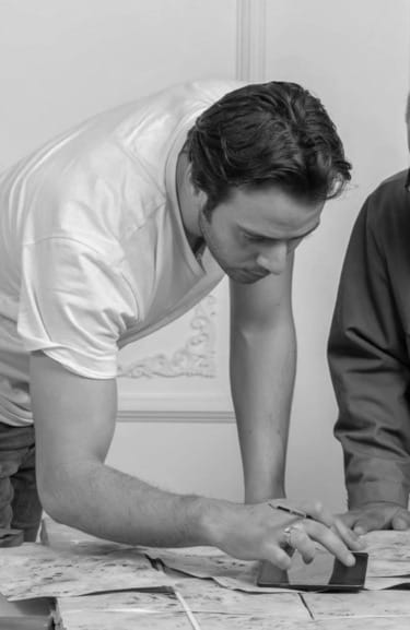 Man in a white t-shirt leaning over a table, using a digital tablet, focusing intently on the screen.