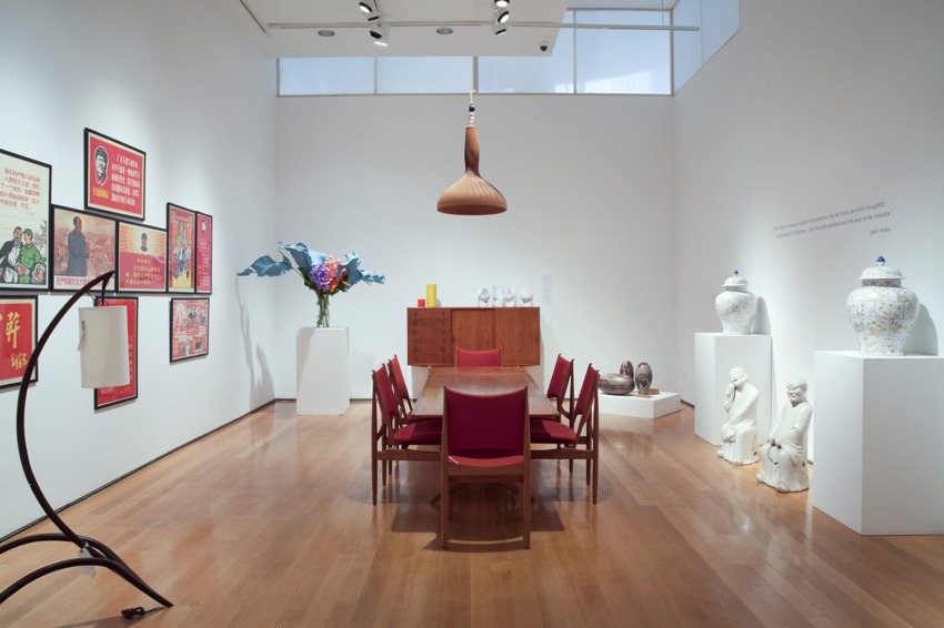 Modern art gallery with vintage posters, sculptures, and a wooden dining table with red chairs and hanging lamp.