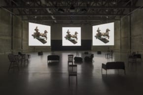 Art installation with two large screens displaying classical sculptures, surrounded by empty chairs in a dimly lit room.