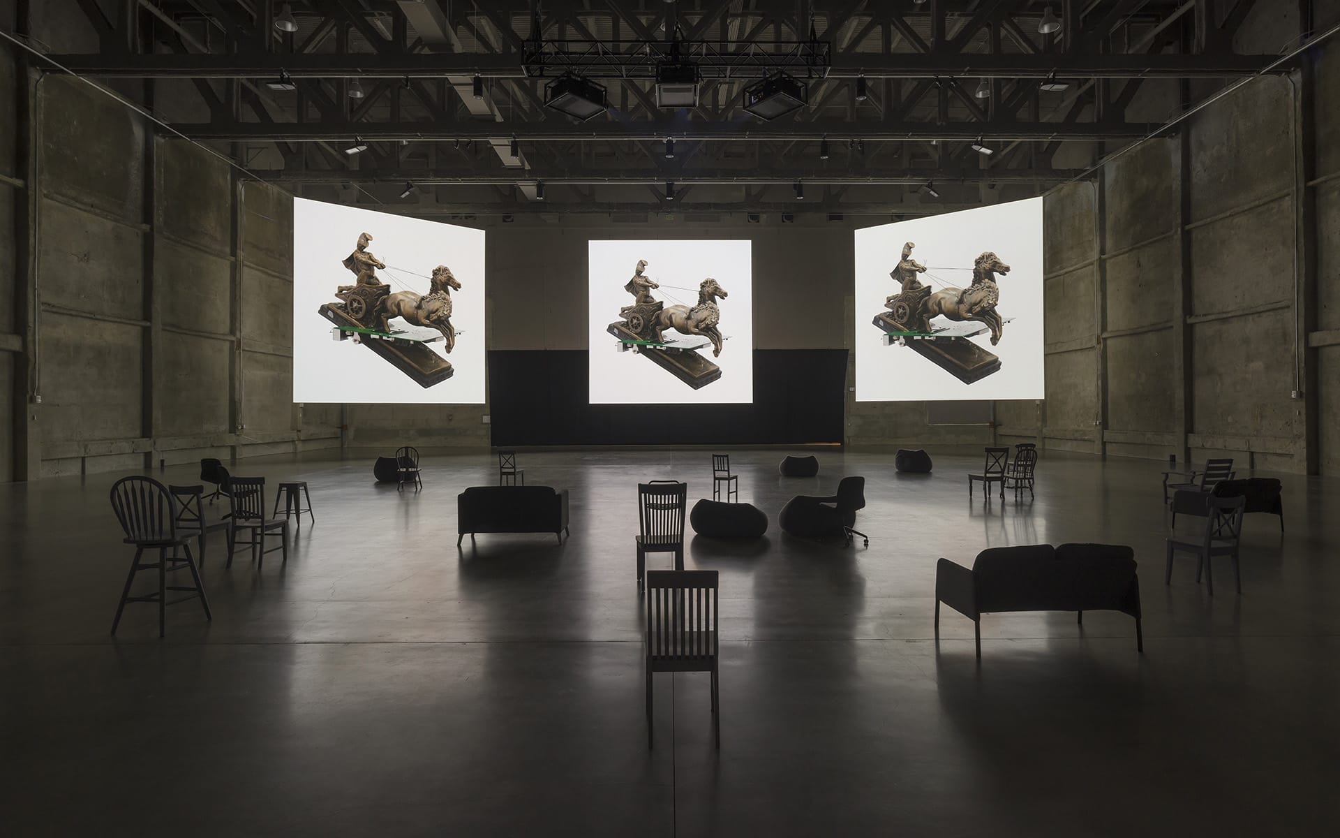 Art installation with two large screens displaying classical sculptures, surrounded by empty chairs in a dimly lit room.