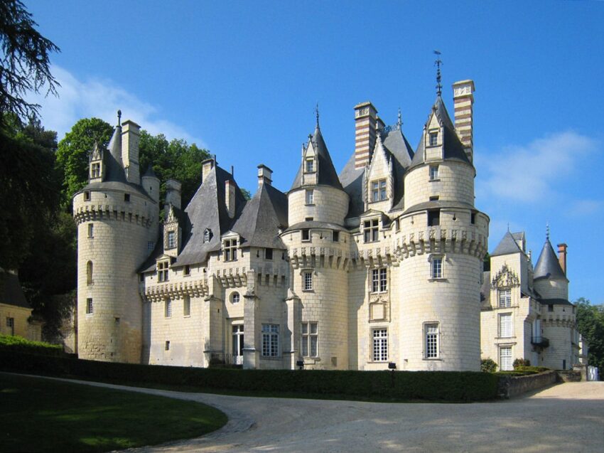 Medieval stone castle with tall turrets and conical roofs, surrounded by trees and set against a clear blue sky.