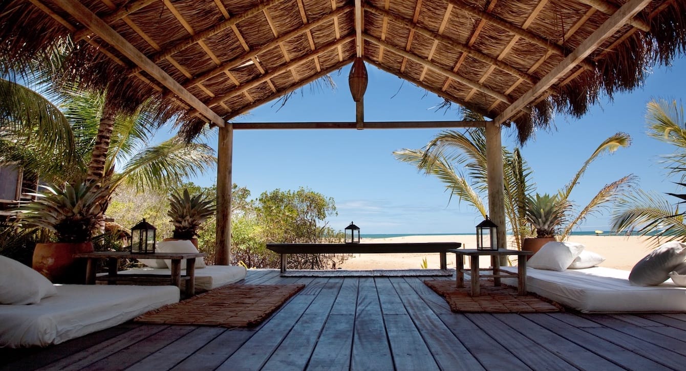 Beach cabana with wooden floor and palm roof, surrounded by tropical plants, overlooking the ocean on a sunny day.