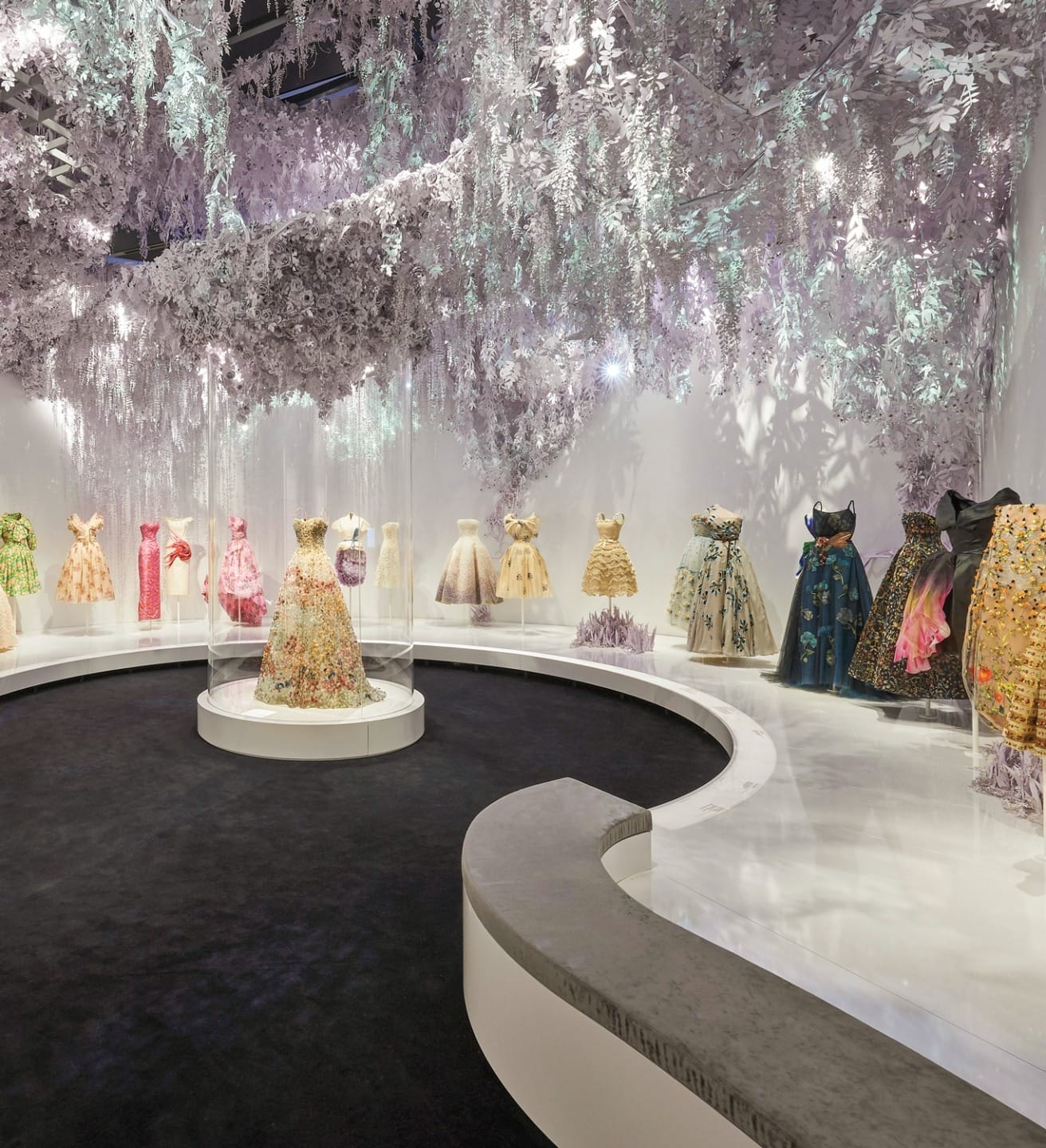 Exhibit of decorative dresses displayed under hanging floral arrangements in a fashion museum setting.
