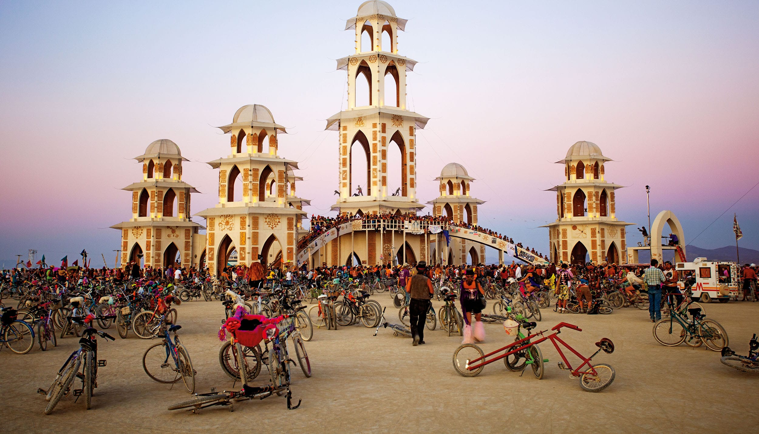 A crowd gathers around a uniquely designed temple with bicycles at sunset in a desert setting.
