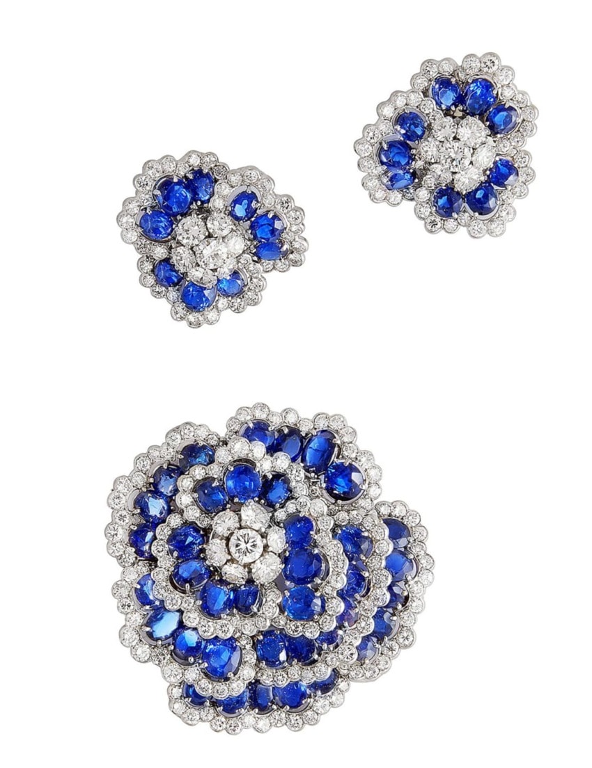 Blue sapphire and diamond floral brooch and earrings set with intricate detailing and sparkling gemstones on a white background.