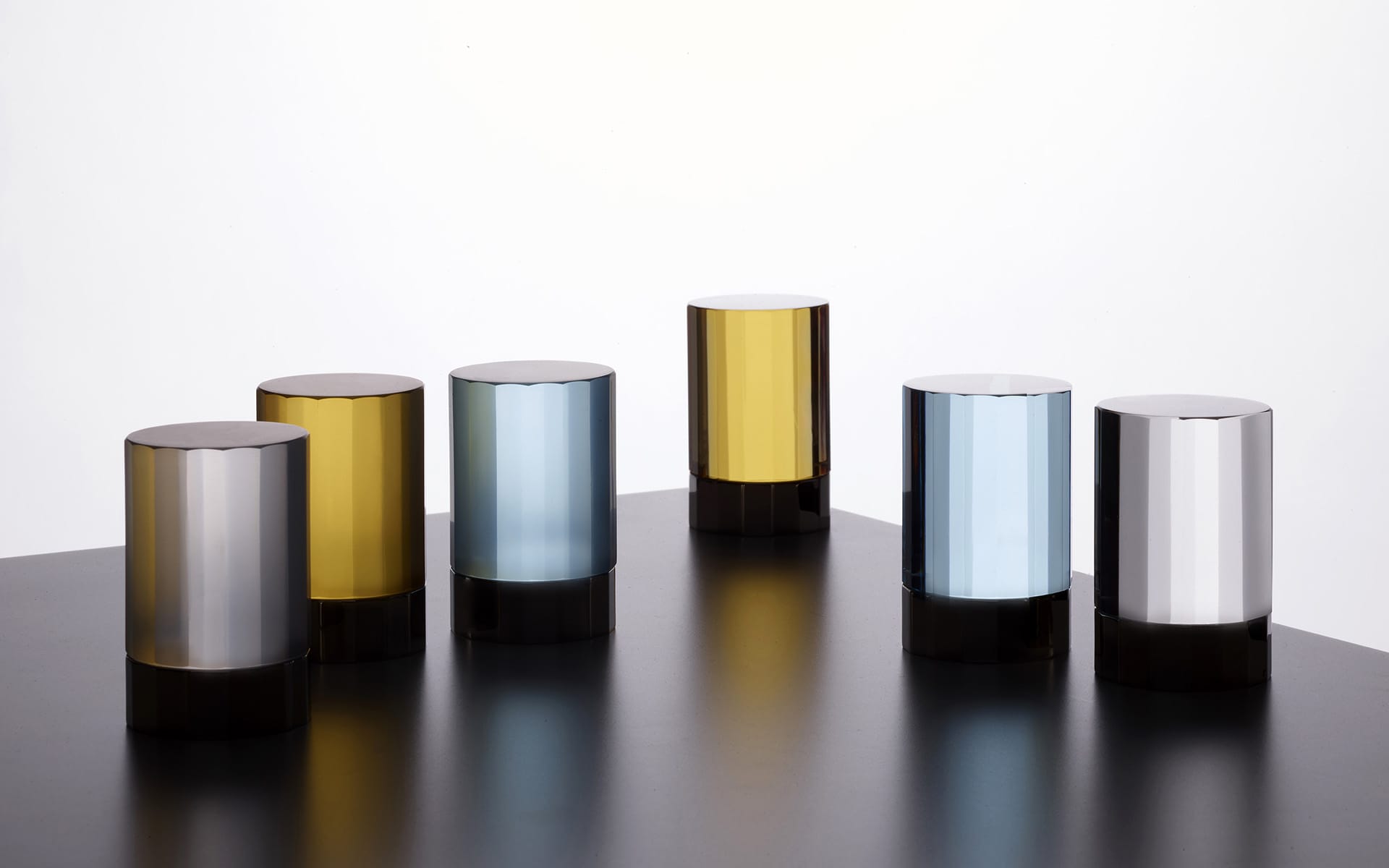 Six cylindrical glass objects in different shades on a dark surface against a white background.