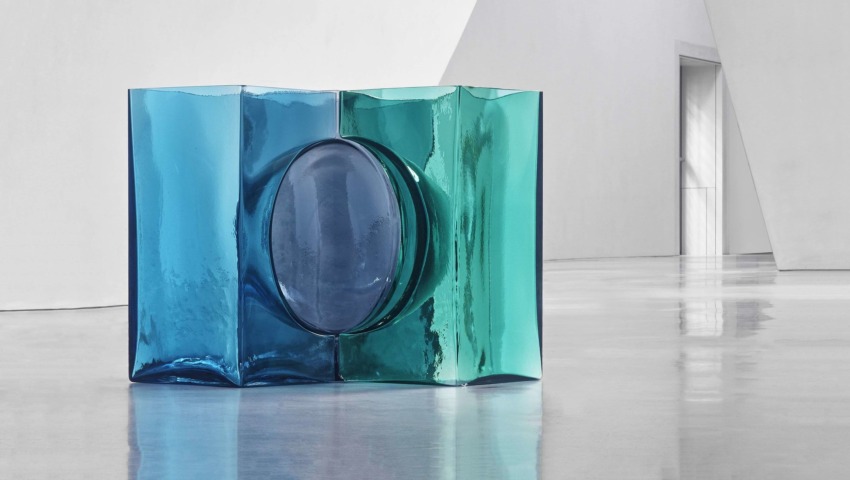 Glass sculpture with intersecting blue and green sections set on a reflective floor in a minimalist white gallery space.
