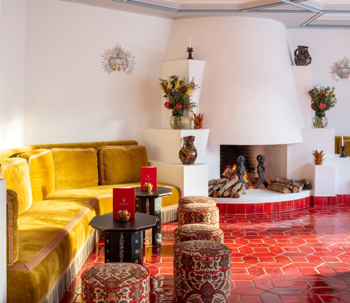 Colorful room with yellow velvet sofa, patterned stools, vibrant flowers, and a corner fireplace on red-tiled floor.