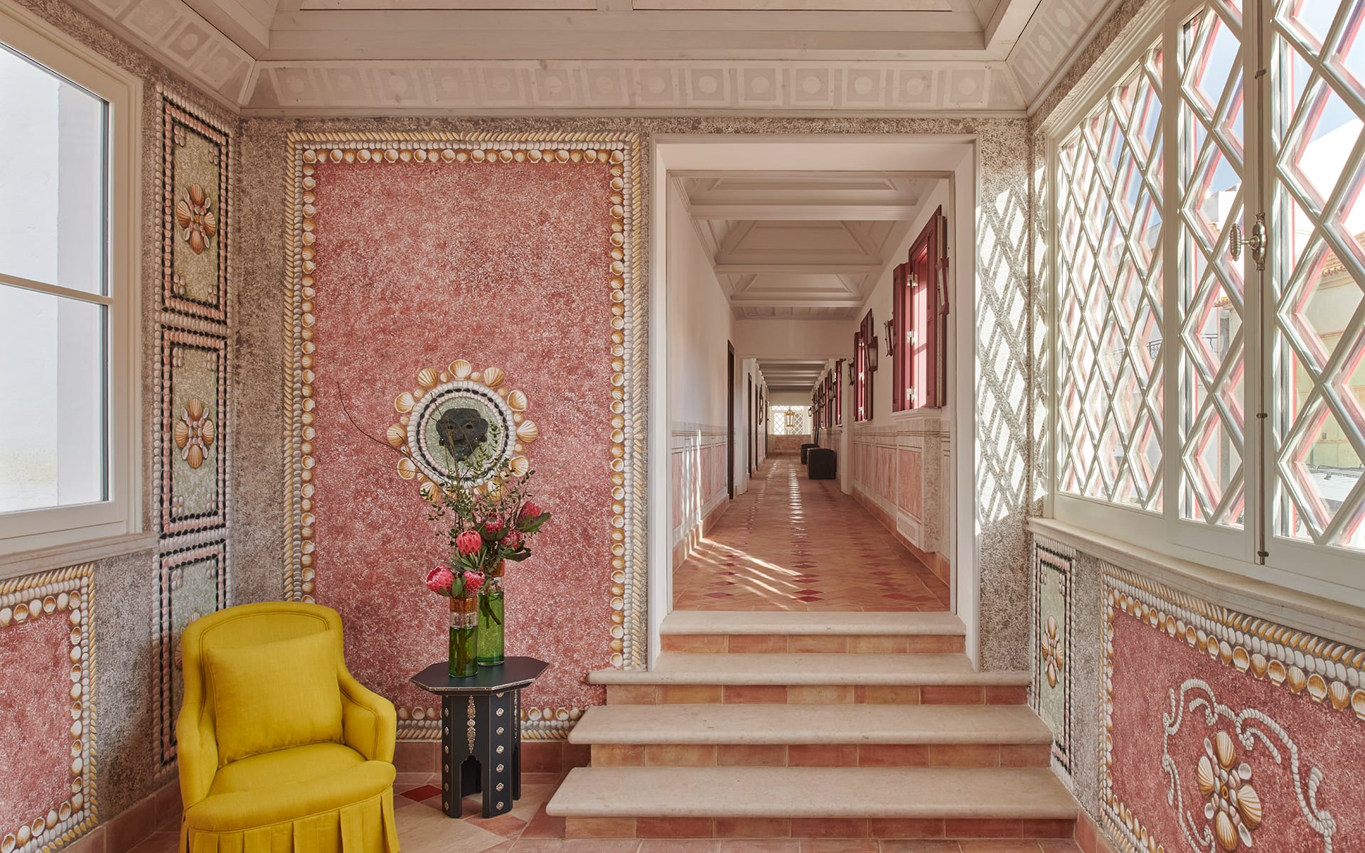 Interior hallway with ornate red and white walls, a yellow chair, a floral vase on a table, and sunlight through lattice windows.