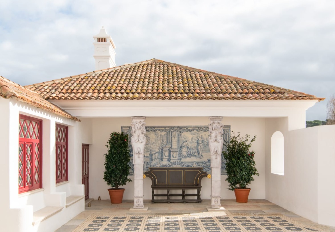 Traditional courtyard with a tiled roof, decorative tile mural, and potted plants next to a wooden bench.