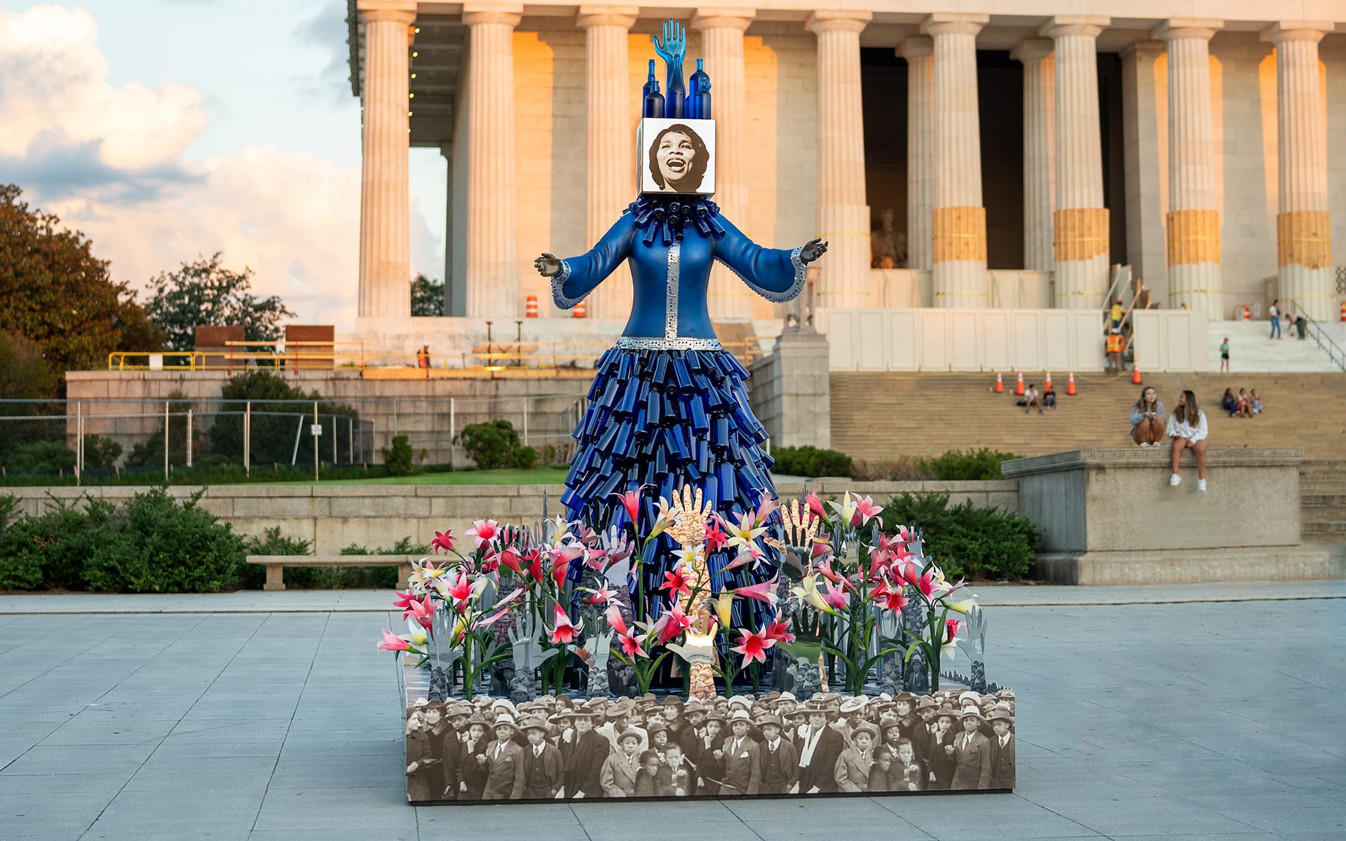 Colorful sculpture in front of a large building, featuring blue and pink elements, surrounded by flowers and historical images.