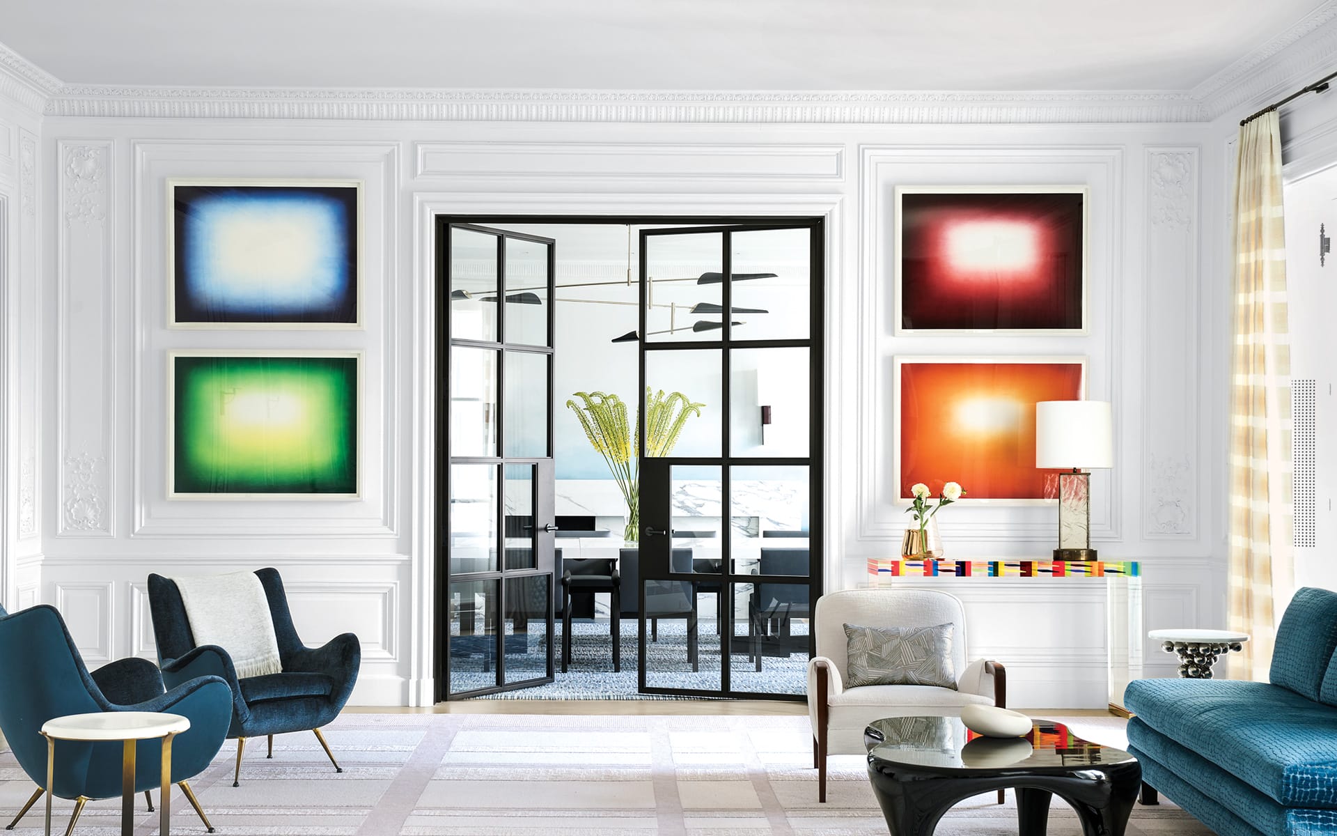 Modern living room with colorful abstract art on walls, elegant furniture, glass doors, and a vase with tall flowers.