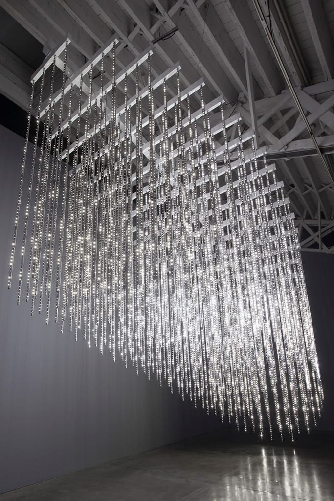 Modern art installation with cascading LED light strips suspended from the ceiling in an industrial-style gallery setting.