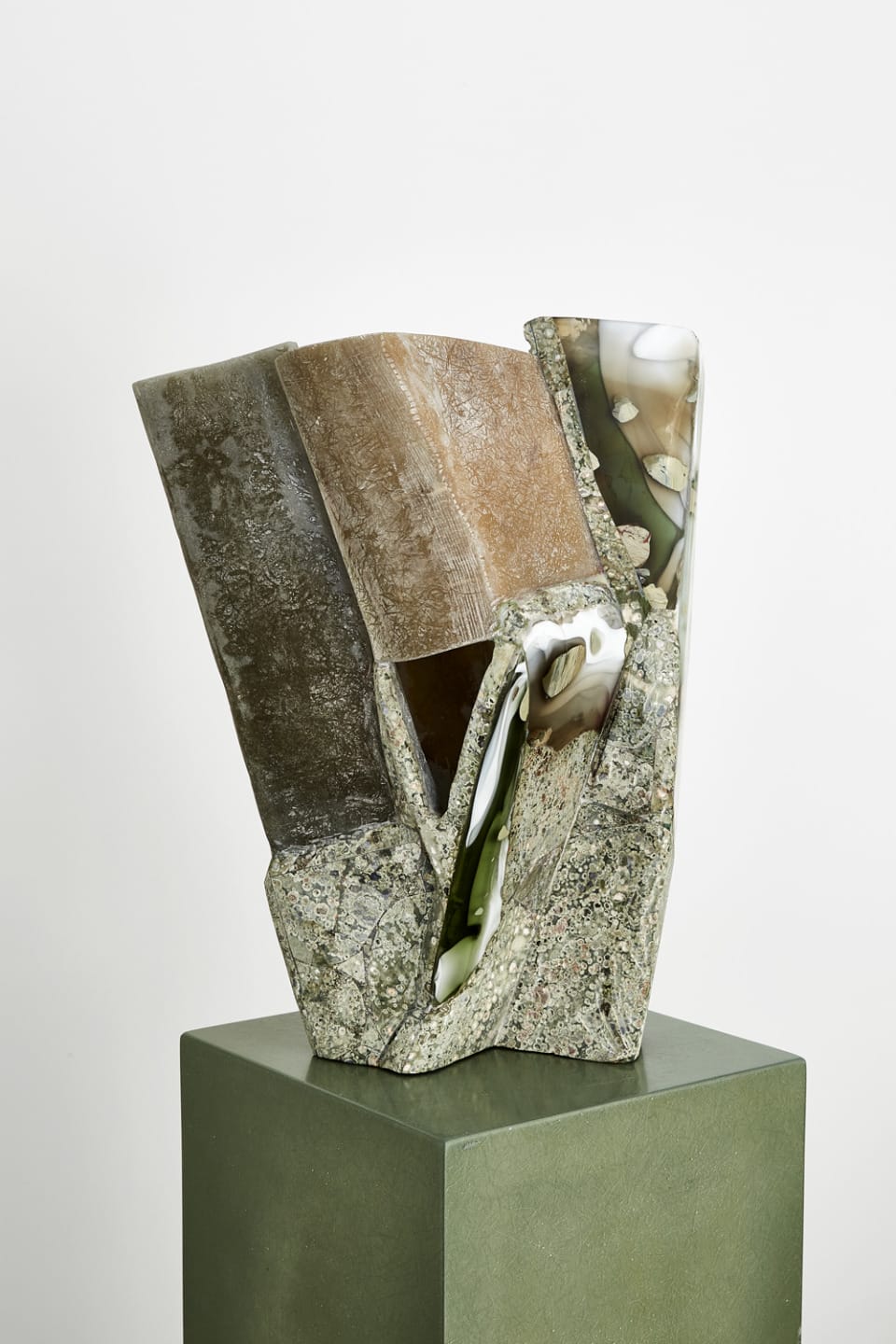 Abstract stone sculpture with geometric shapes and varied textures on a green pedestal against a plain background.