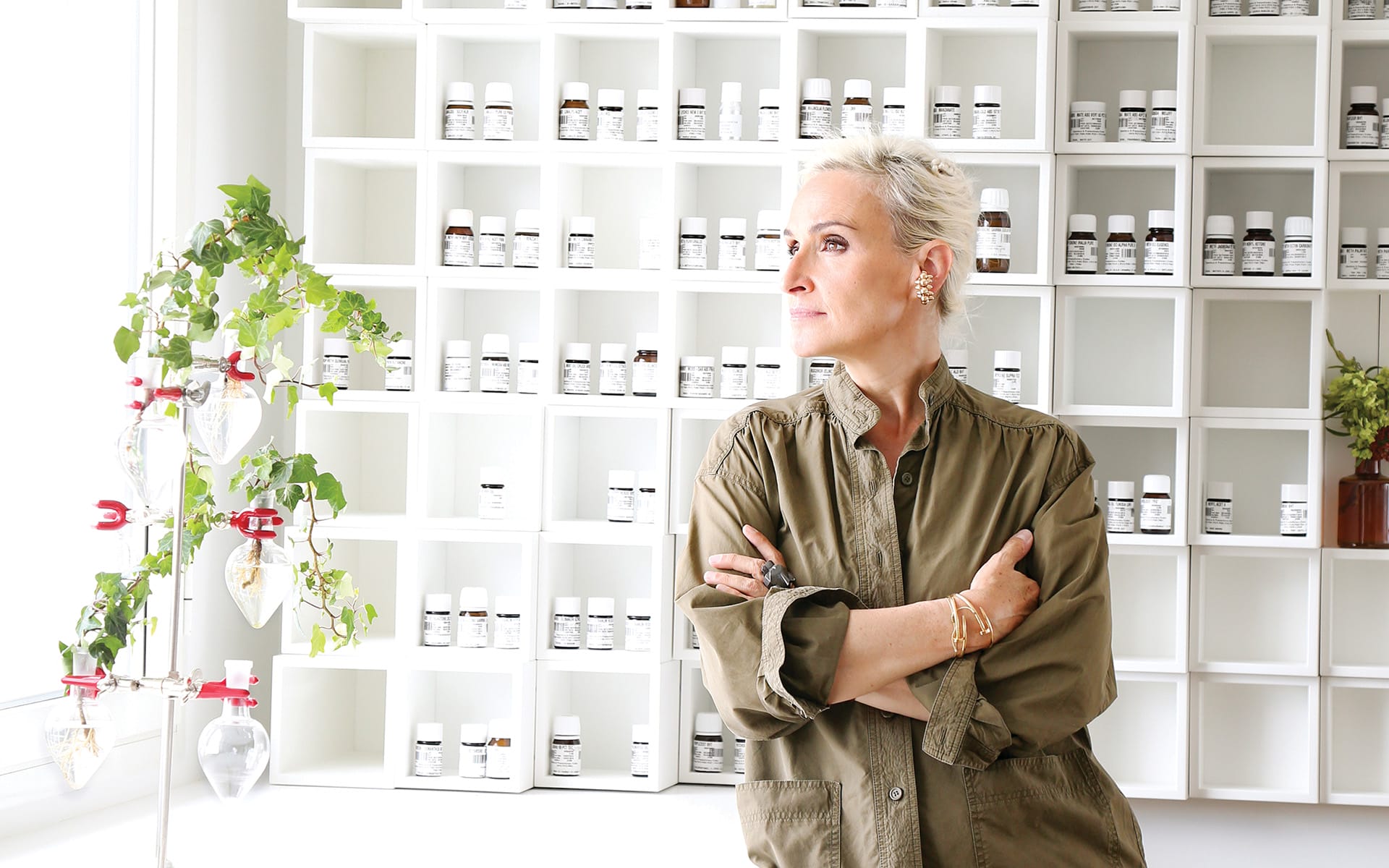 Person in a green shirt standing in front of a wall with small bottles on shelves and a plant on the side.