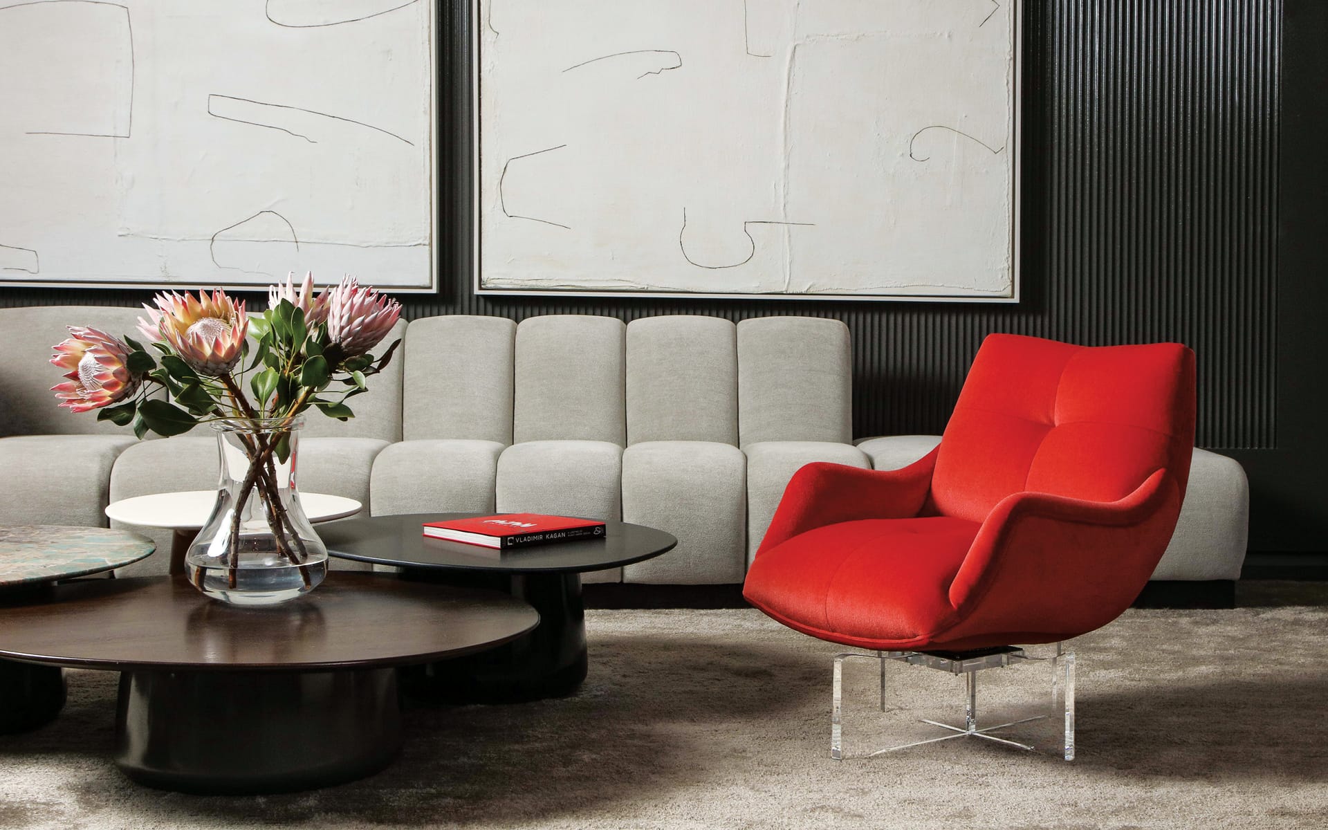 Modern living room with gray sofa, vibrant red armchair, two glass vases with flowers, and abstract art on the wall.