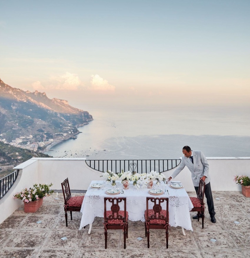 Server arranges table set for dinner on a terrace overlooking a coastal view at sunset.