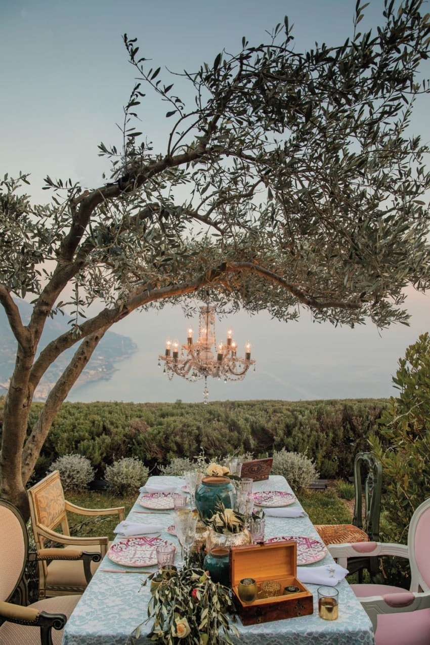 Outdoor dining table set under tree with chandelier, overlooking a scenic landscape with sea and hills in the background.