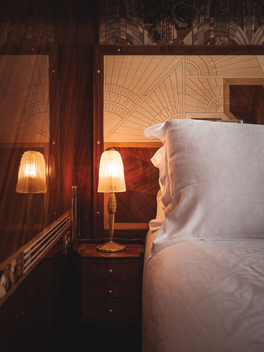 Art Deco style hotel room with a lit bedside lamp, ornate wooden headboard, and crisp white bedding.