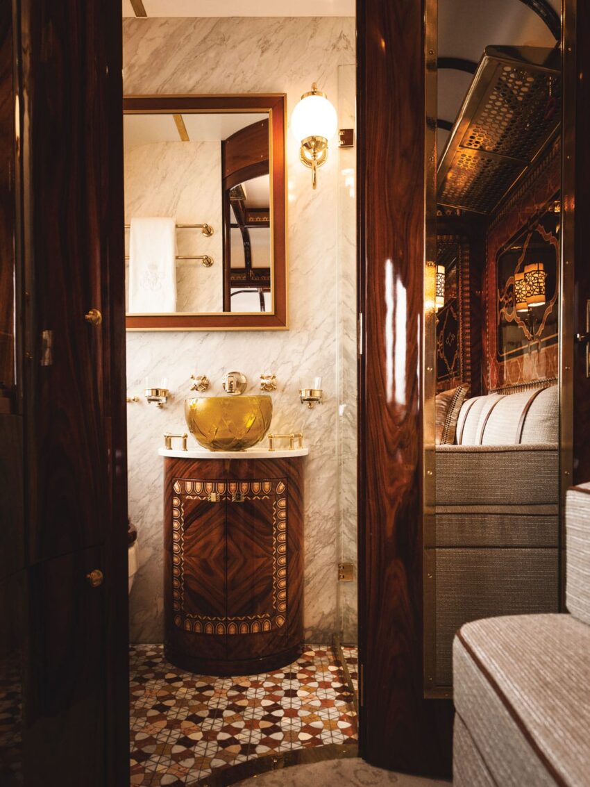 Luxury train bathroom with ornate woodwork, marble walls, a gold sink, and mosaic tile floor, reflecting opulent design.