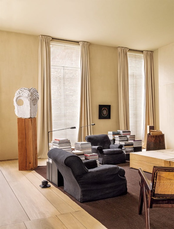 Cozy living room with black armchairs, tall windows, and stacks of books, featuring a sculpture and wooden table.