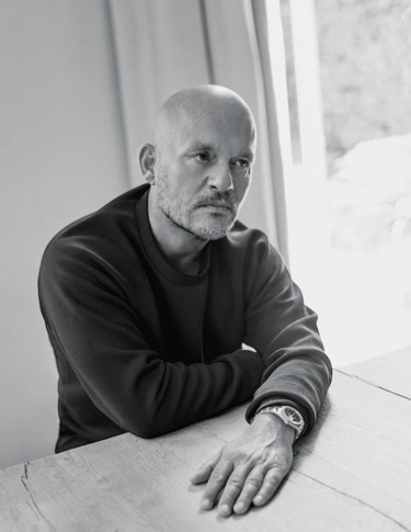Man seated at a table wearing a dark sweater and a watch, looking contemplative in a softly lit room.