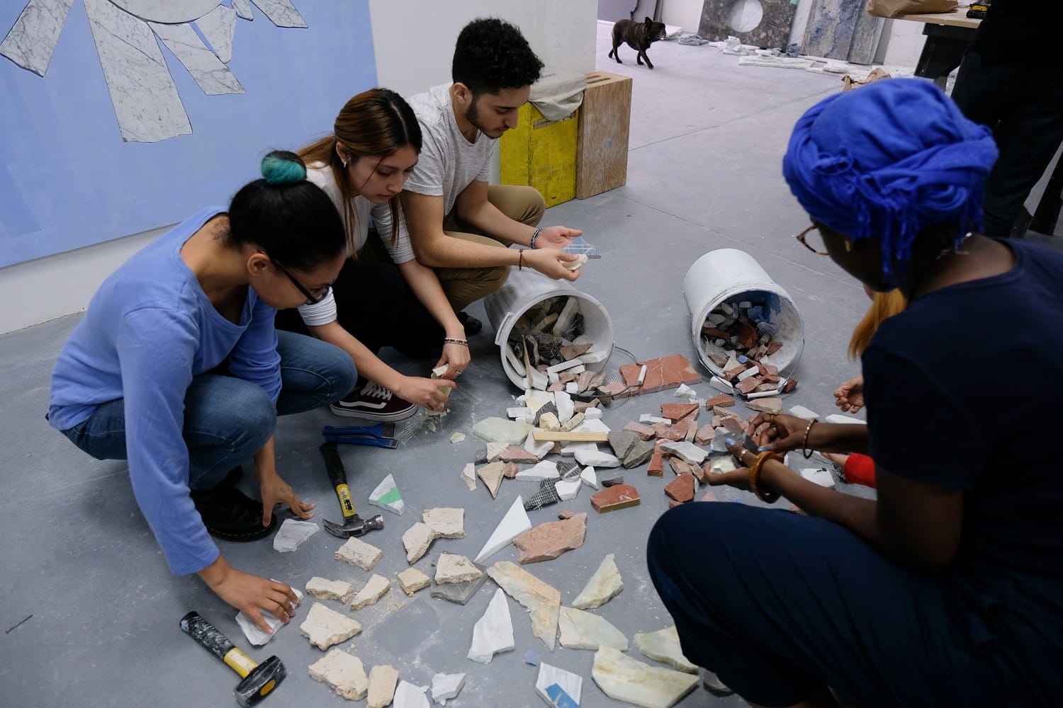 Four people sorting tiles and rocks from spilled buckets on the floor in an art studio setting.