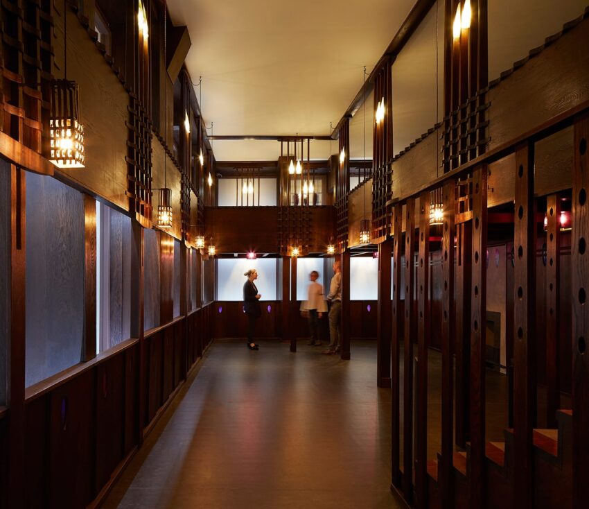 Interior view of a dimly lit, art deco-style room with people standing, featuring wood paneling and geometric lights.