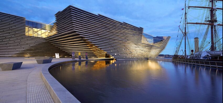 Modern architectural building beside a calm body of water with a historic ship and benches in the foreground, under a blue sky.