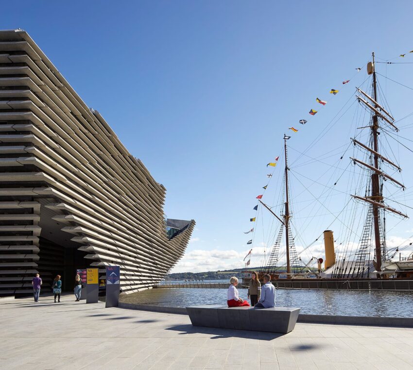 Museum with modern architecture next to an old sailing ship on a sunny day, people sitting on a bench by the water.