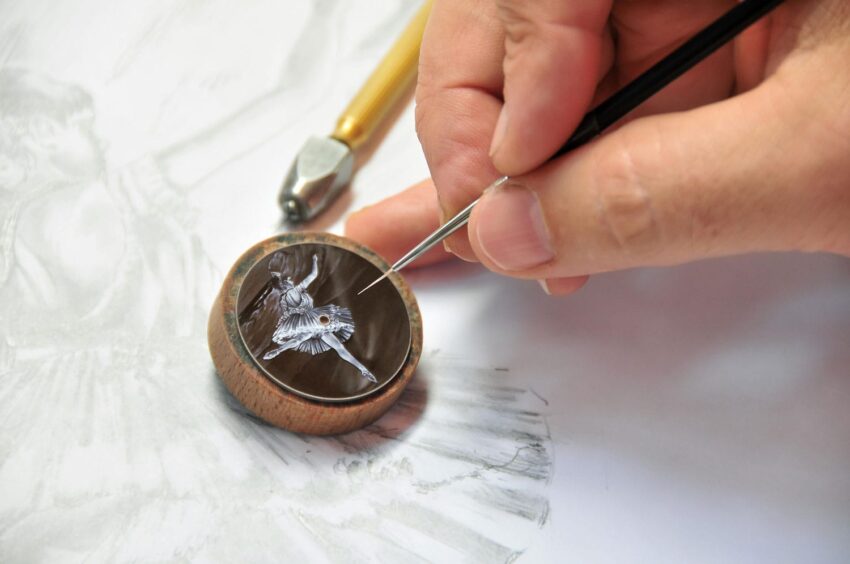 Hand painting a detailed miniature sculpture inside a ring-like setting, with drawing tools on paper in the background.