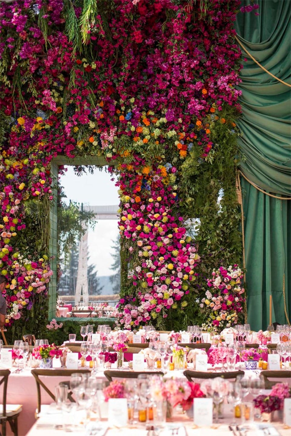 Lavish wedding venue with vibrant floral wall, draped greenery, elegant table settings, and colorful flowers.