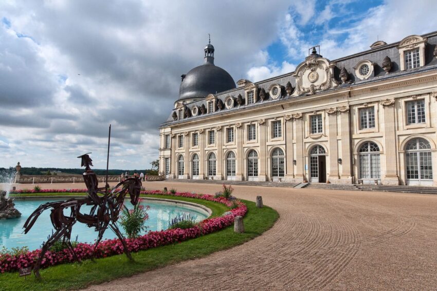 Historic chateau with a domed roof, ornate architecture, and a metal sculpture of a rider near a circular fountain with flowers.