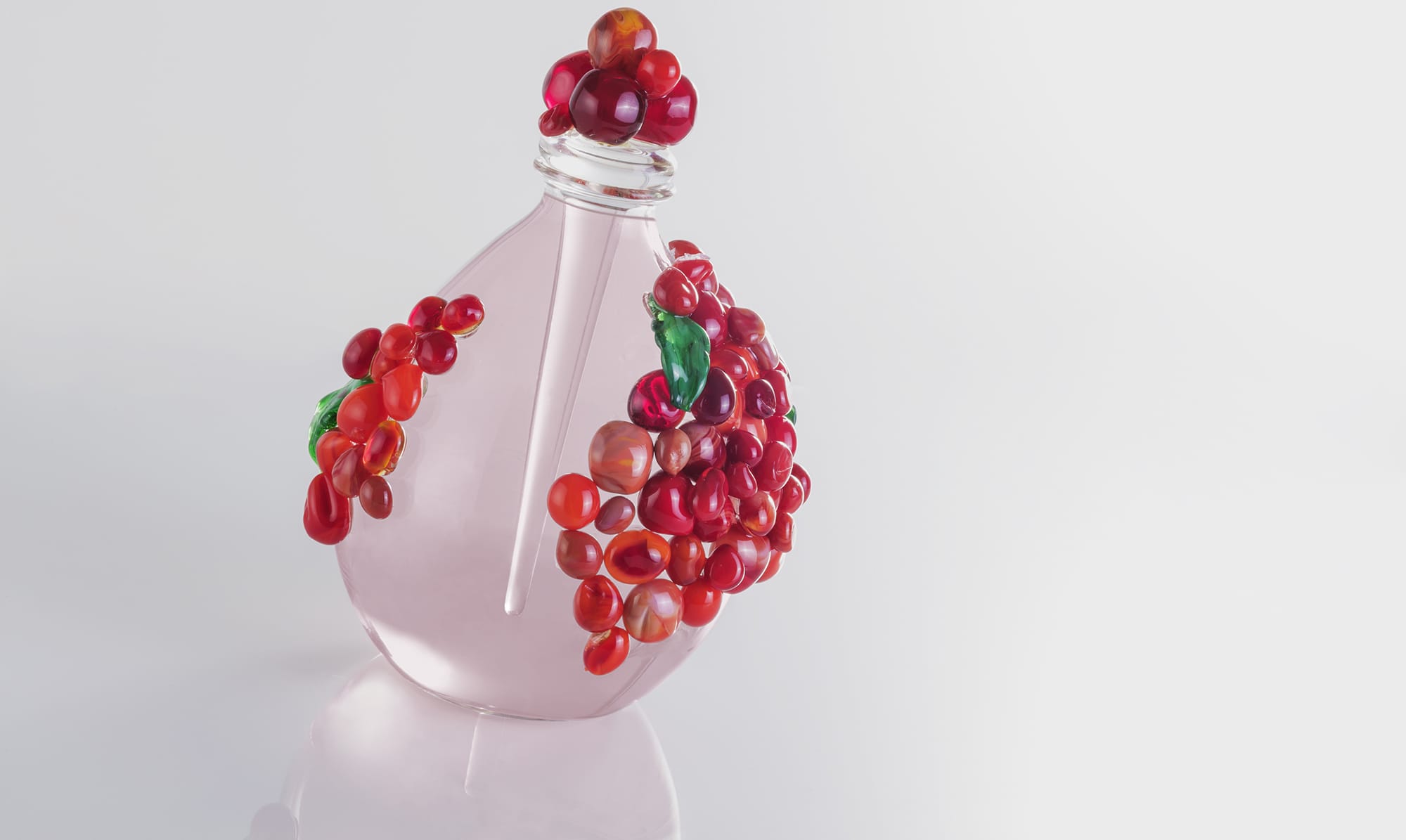 Elegant glass bottle adorned with clusters of red and pink glass beads, reflecting light on a simple white background.